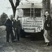 Trabajadores de la imprenta Quintilla y Cardona S.L. de Barcelona celebrando el Día del Santo Patrón de las Artes Gráficas el 6 de mayo, frente al autobús de la empresa con pancarta, años cincuenta