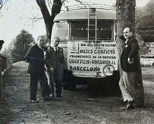 Trabajadores de la imprenta Quintilla y Cardona S.L. de Barcelona celebrando el Día del Santo Patrón de las Artes Gráficas el 6 de mayo, frente al autobús de la empresa con pancarta, años cincuenta